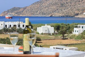 A coastal view with a boat on the sea, white buildings, and a table with a wine bottle and glass. - Sifnaika Konakia ~ Armada Residence (Sifnos)