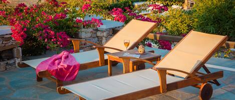 Two wooden lounge chairs with white cushions, a small table, and a glass of drink on a stone patio with pink bougainvillea flowers.