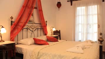 A neatly made bed with white linens and red pillows, a bedside table with a lamp, and a window with sheer curtains.
