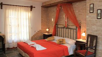 Un dormitorio con pared de ladrillo, una cama con colcha roja, dos mesitas de noche con lámparas, una silla de madera y una ventana con cortinas traslúcidas.