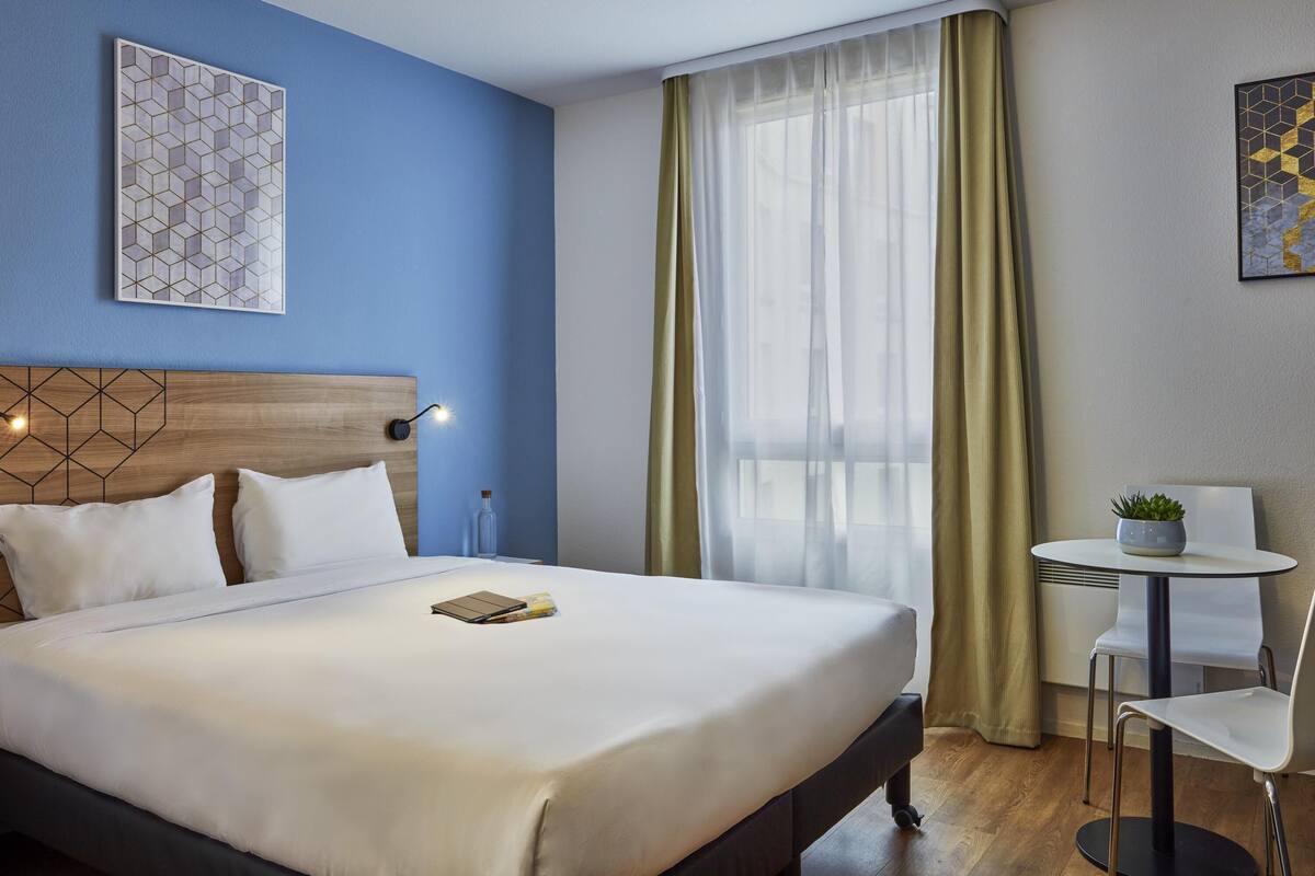 Ett hotellrum med en säng, ett sängbord med en lampa, ett litet bord med en stol och ett fönster med gardiner.