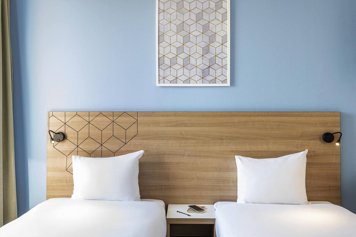 Ein Hotelzimmer mit hölzernem Kopfteil, zwei Betten mit weißer Bettwäsche, einem Nachttisch mit Lampe und einem Bilderrahmen mit geometrischem Kunstwerk an der Wand.