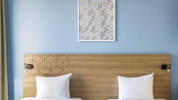 Una habitación de hotel con cabecera de madera, dos camas con ropa blanca, una mesita de noche con lámpara y un cuadro abstracto geométrico en la pared.