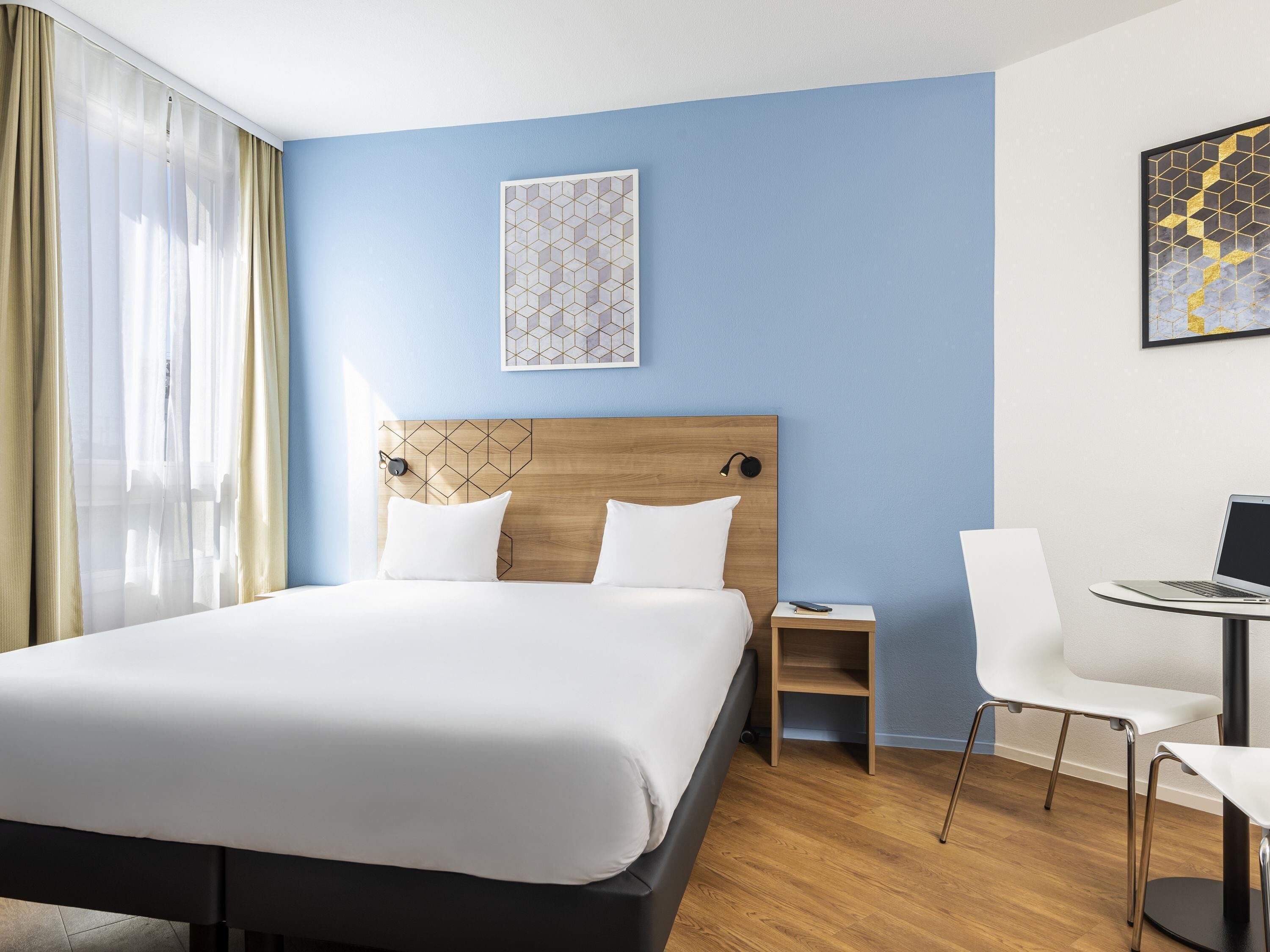 A hotel room with a bed, bedside table, chair, and a desk with a laptop.