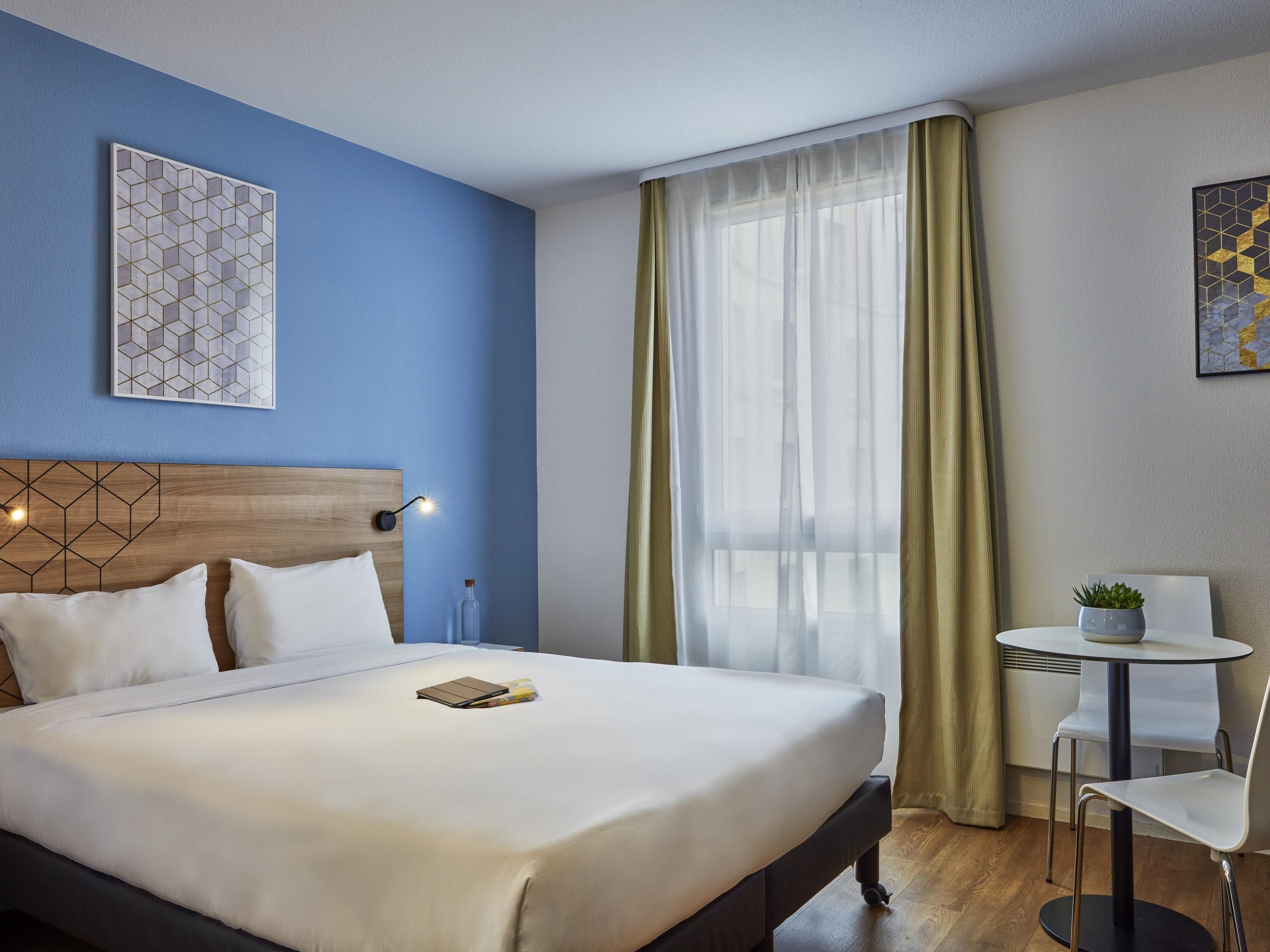 A hotel room with a bed, a bedside table with a lamp, a small table with a chair, and a window with curtains.