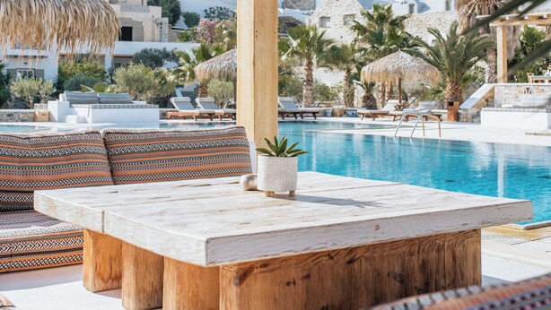 A poolside area with a thatched umbrella, wooden seating, and a view of white buildings in the background.