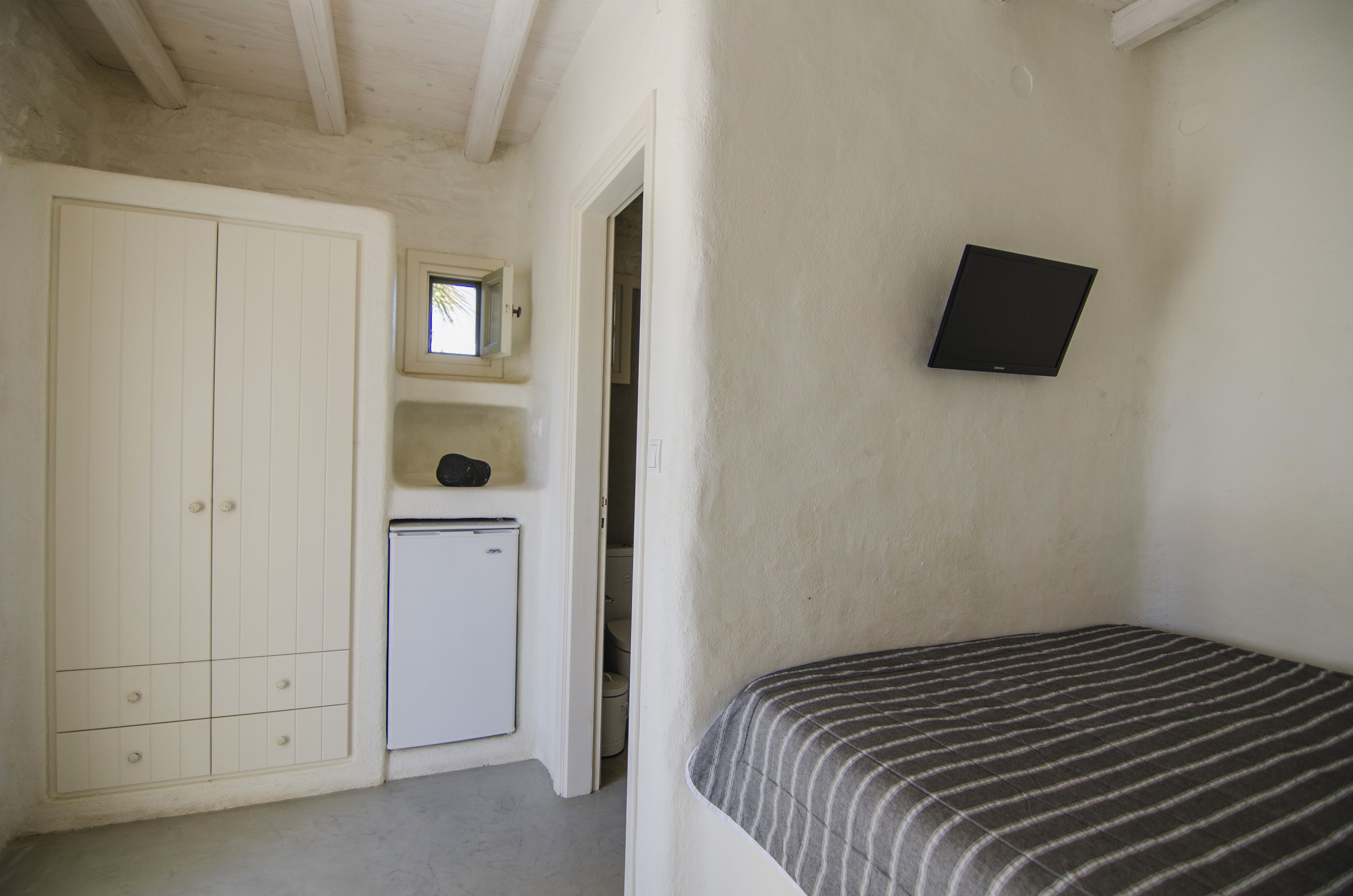 Ein kleiner, schlichter Raum mit einem Bett, einem weißen Schrank, einer kleinen Kühlbox und einem an der Wand befestigten Fernseher.
