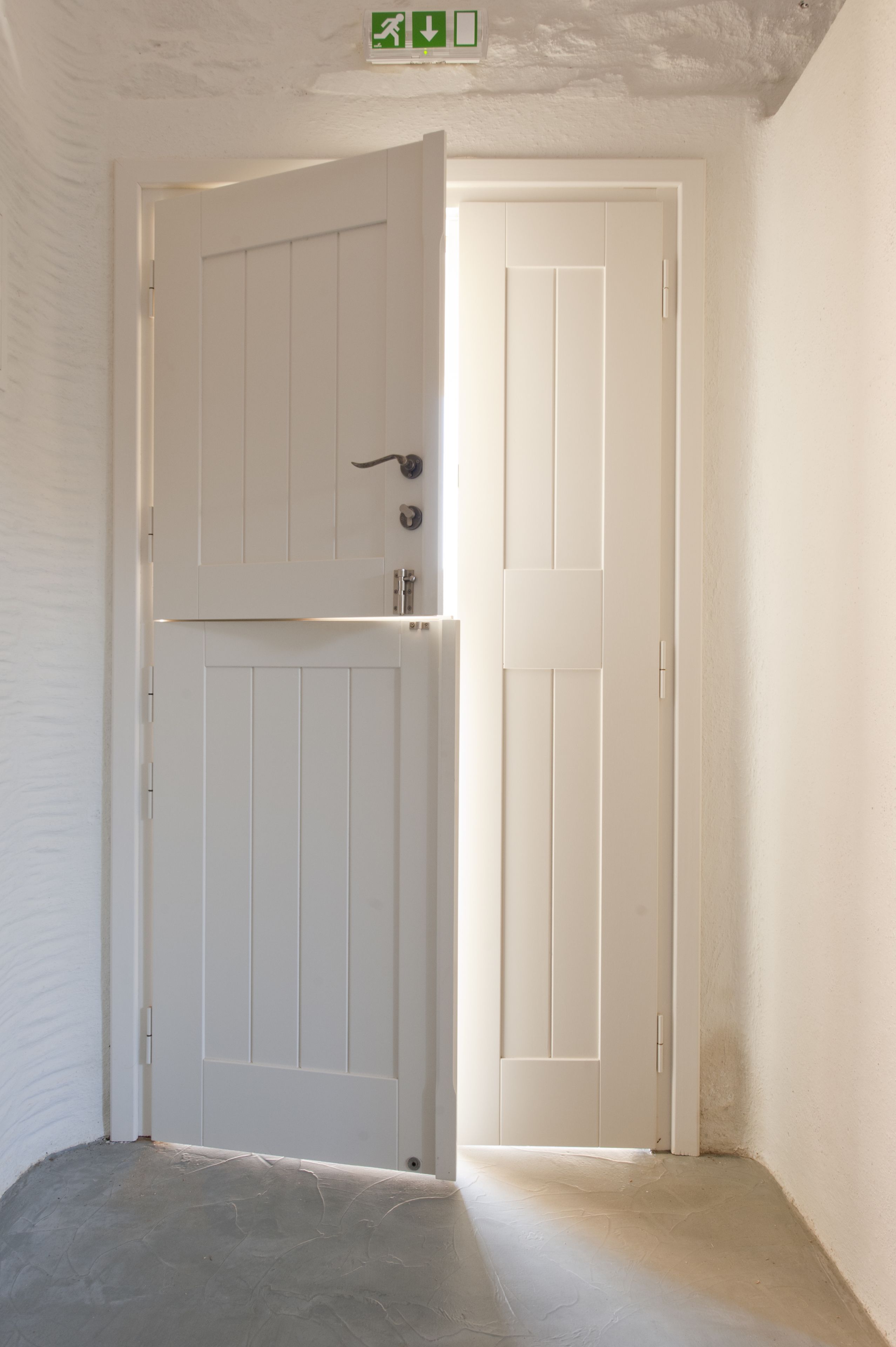 Una porta bianca con maniglia e serratura, che dà accesso a una stanza con pavimento in cemento e pareti bianche.