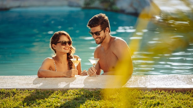 Dos personas sentadas junto a una piscina, con bebidas, rodeadas de follaje.