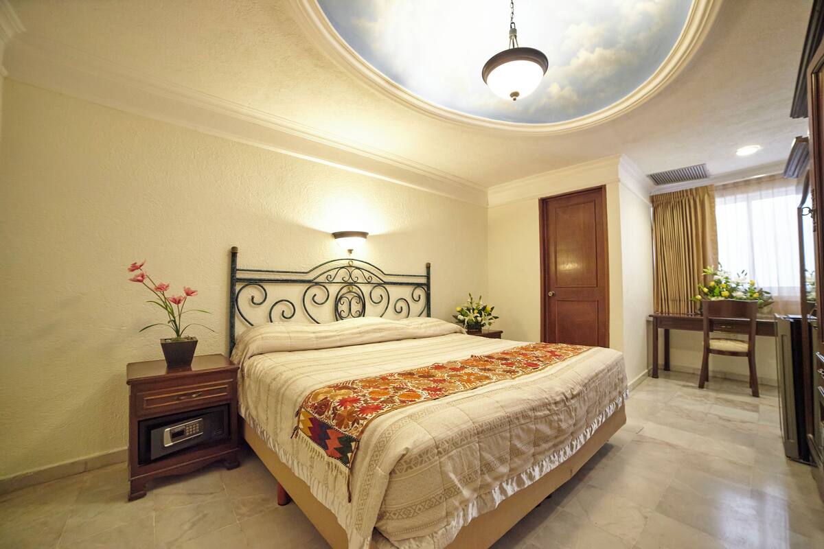 A bedroom with a bed, bedside table, and a ceiling mural.