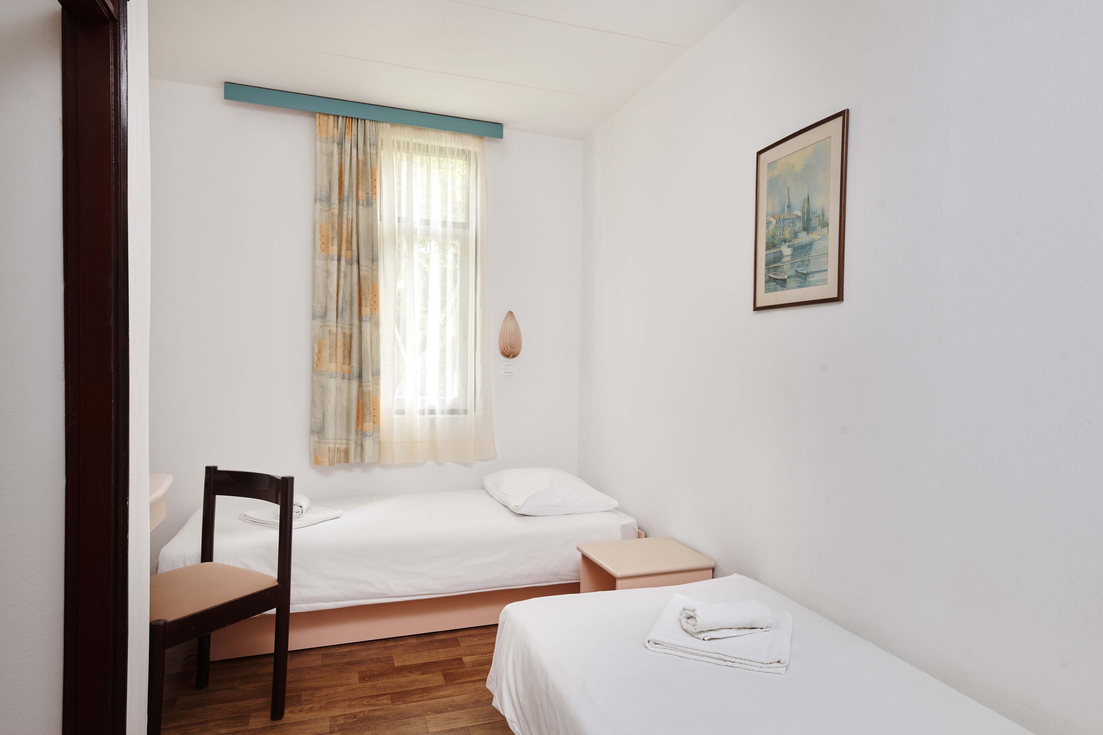 Une petite chambre propre, avec deux lits, une chaise, une table de chevet et une fenêtre ornée de rideaux légers.