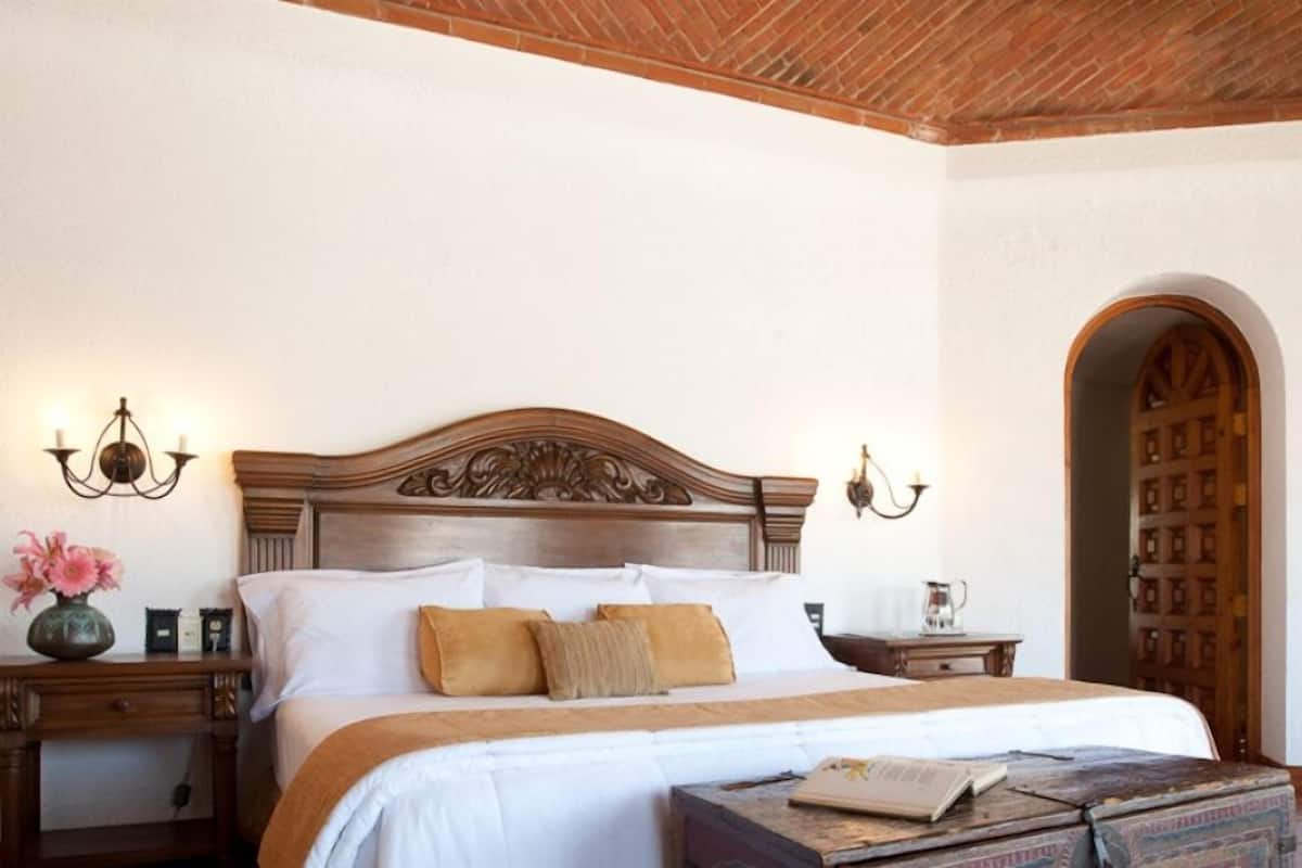 Un dormitorio con cabecera de madera, ropa de cama blanca, una mesita de noche con lámpara y una pequeña mesa con un libro.