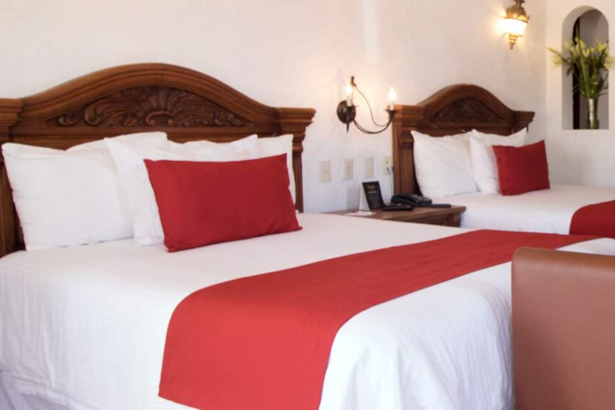 Habitación de hotel con dos camas, cada una con cabecero de madera, ropa de cama blanca y roja, y una mesita de noche con lámpara. También hay una silla de madera y un escritorio con una silla en la habitación.