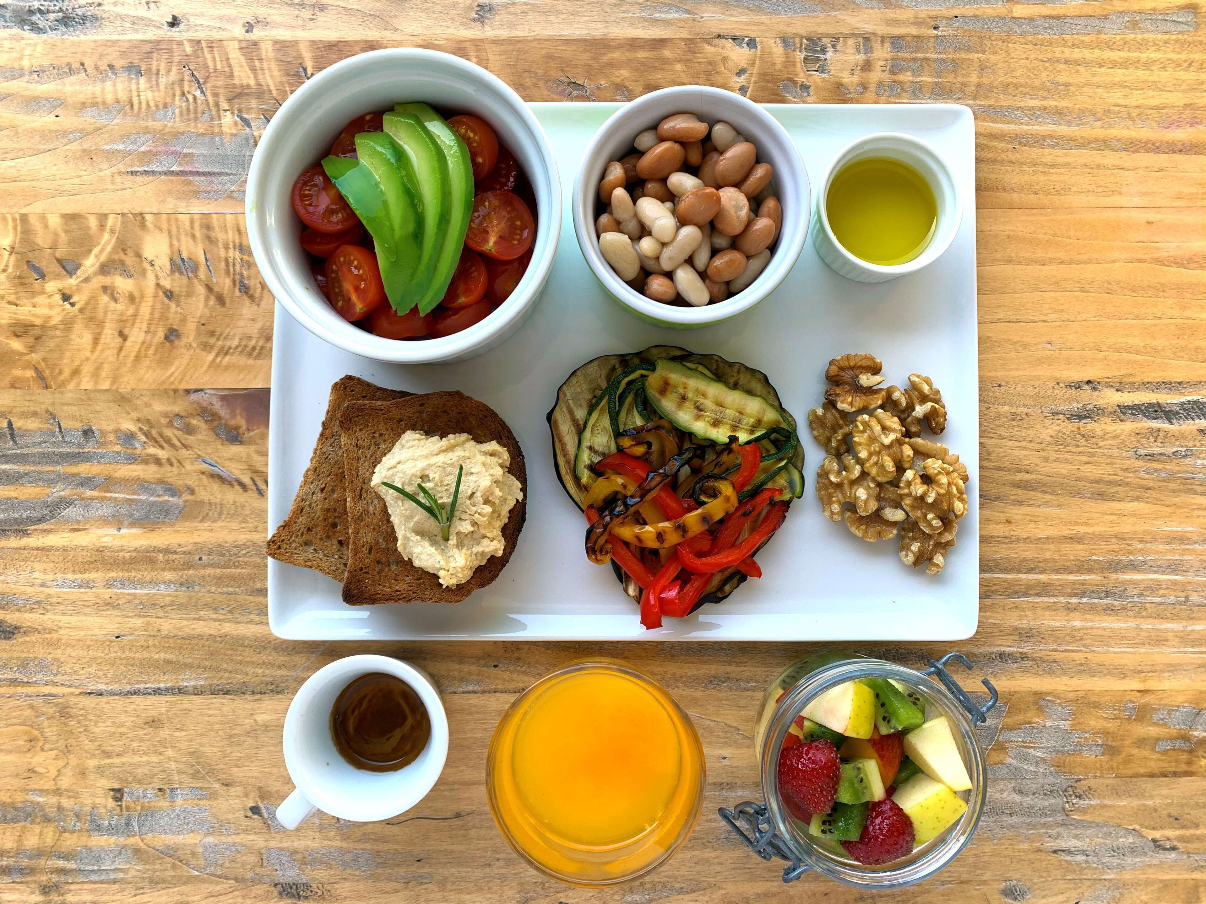 Ein Tablett mit verschiedenen Speisen, darunter Tomaten, Bohnen, Avocado, Walnüsse, gegrilltes Gemüse und ein Glas Orangensaft.