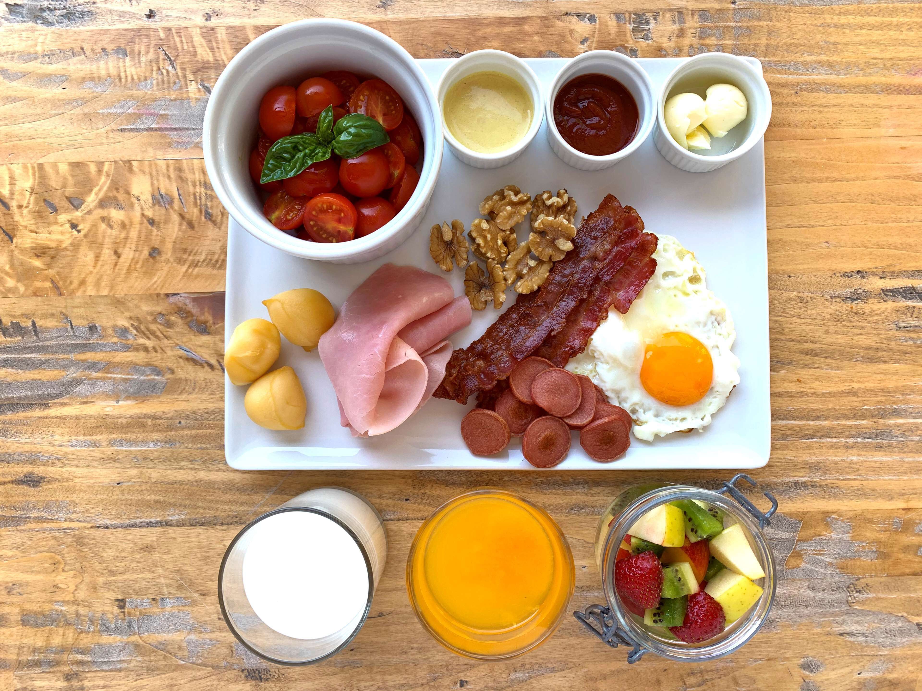 A breakfast tray with eggs, bacon, ham, sausages, tomatoes, walnuts, and a glass of orange juice.