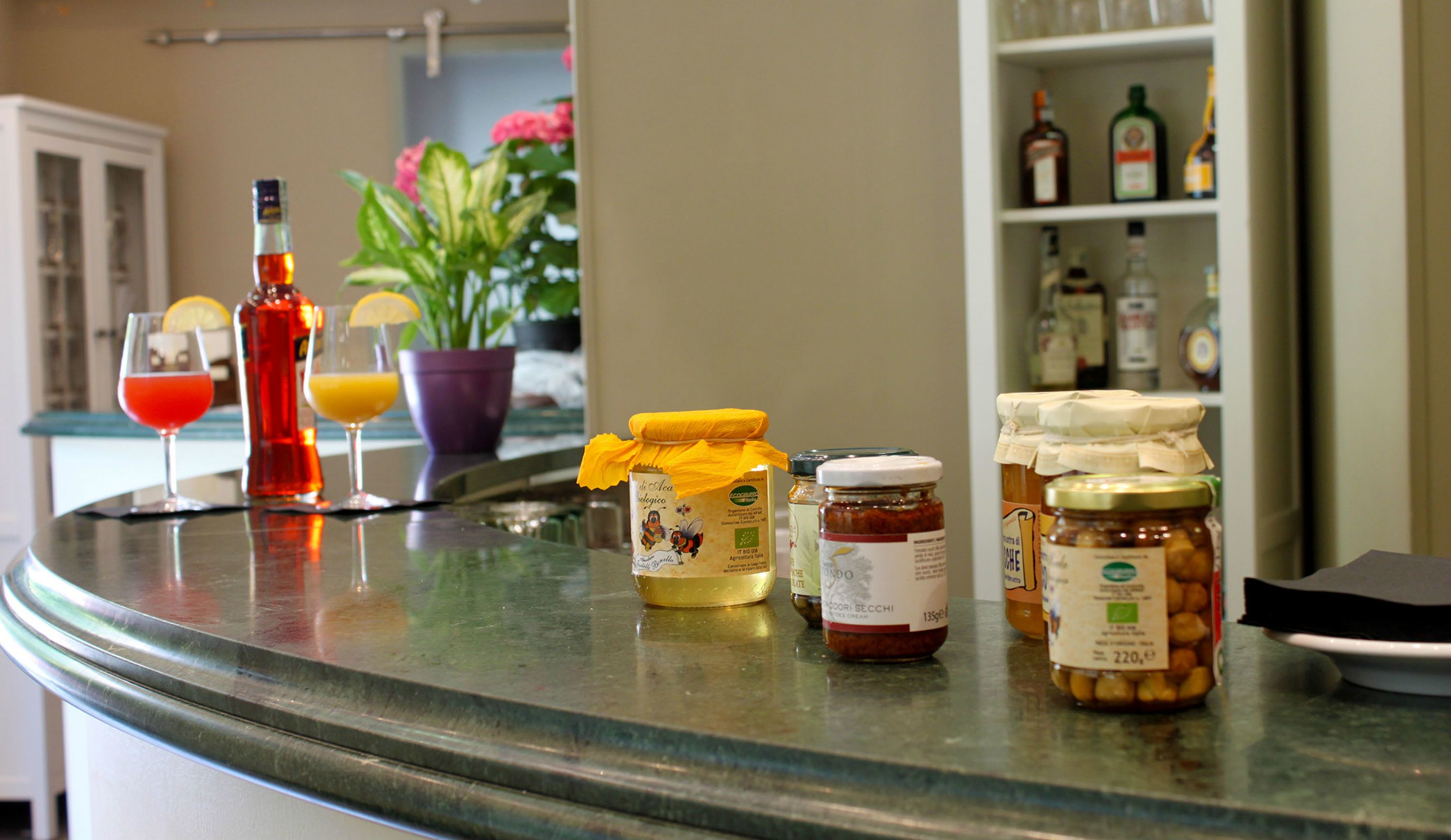 A kitchen counter with various food items, including jars of honey and jam, a bottle of olive oil, and a glass of red wine.