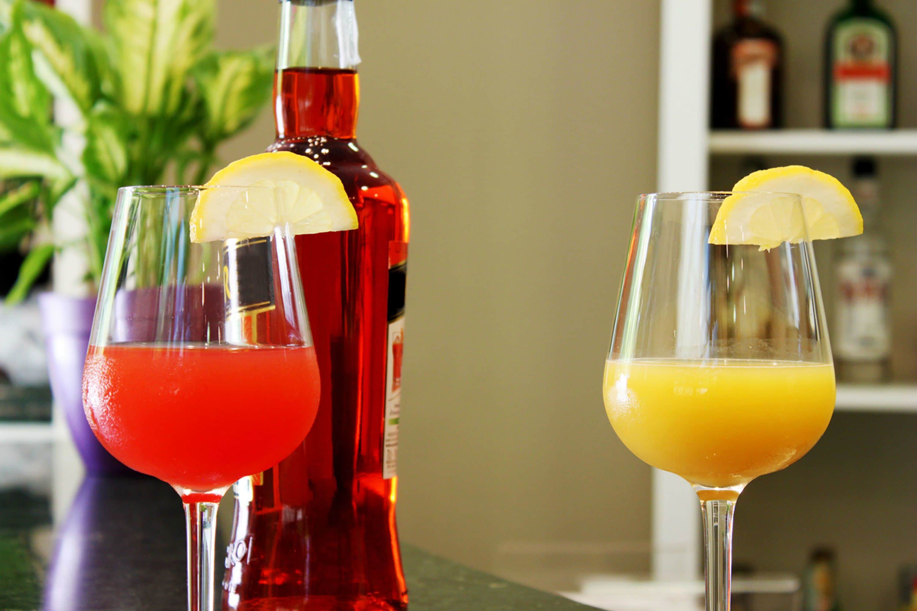 Three glasses of beverages with lemon slices, a bottle of red liquid, and a bottle of orange juice on a counter.