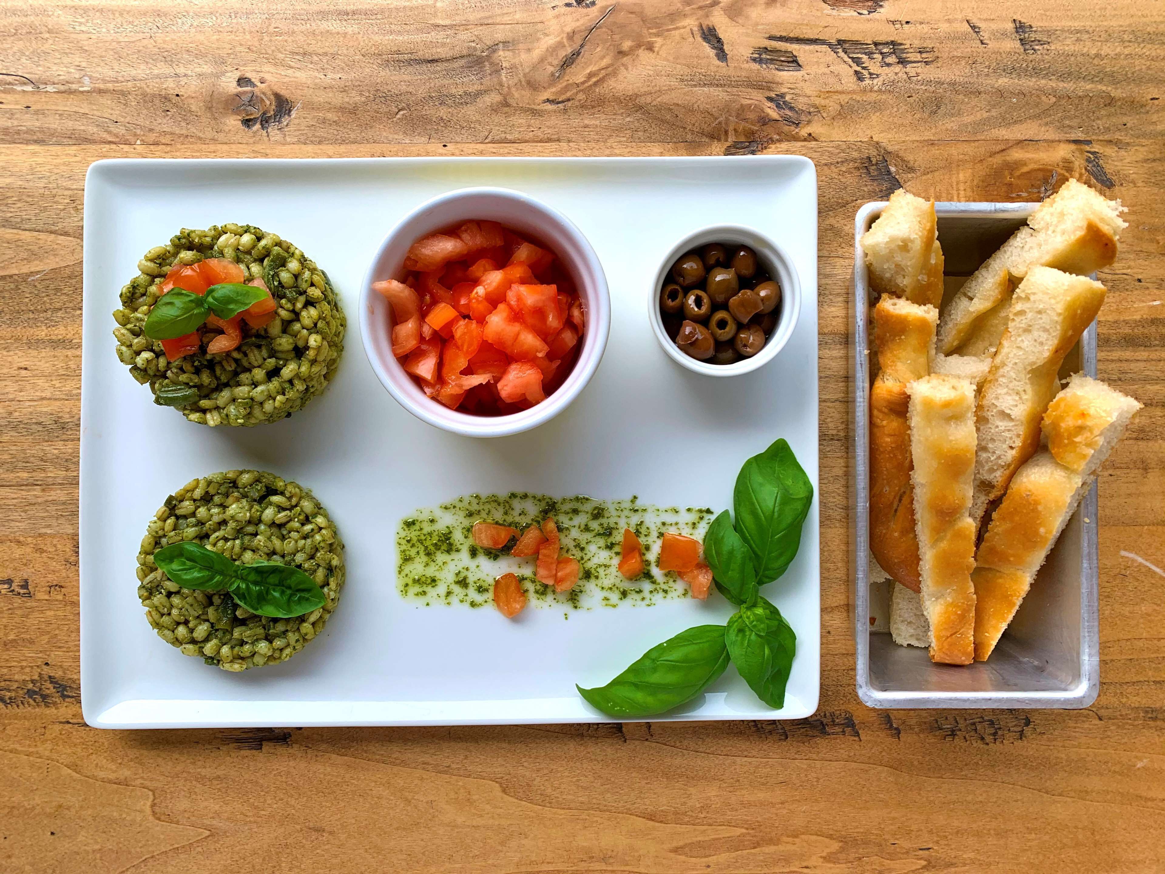 A white rectangular plate with food items including pesto balls, diced tomatoes, olives, and a pesto sauce, accompanied by a container of breadsticks.