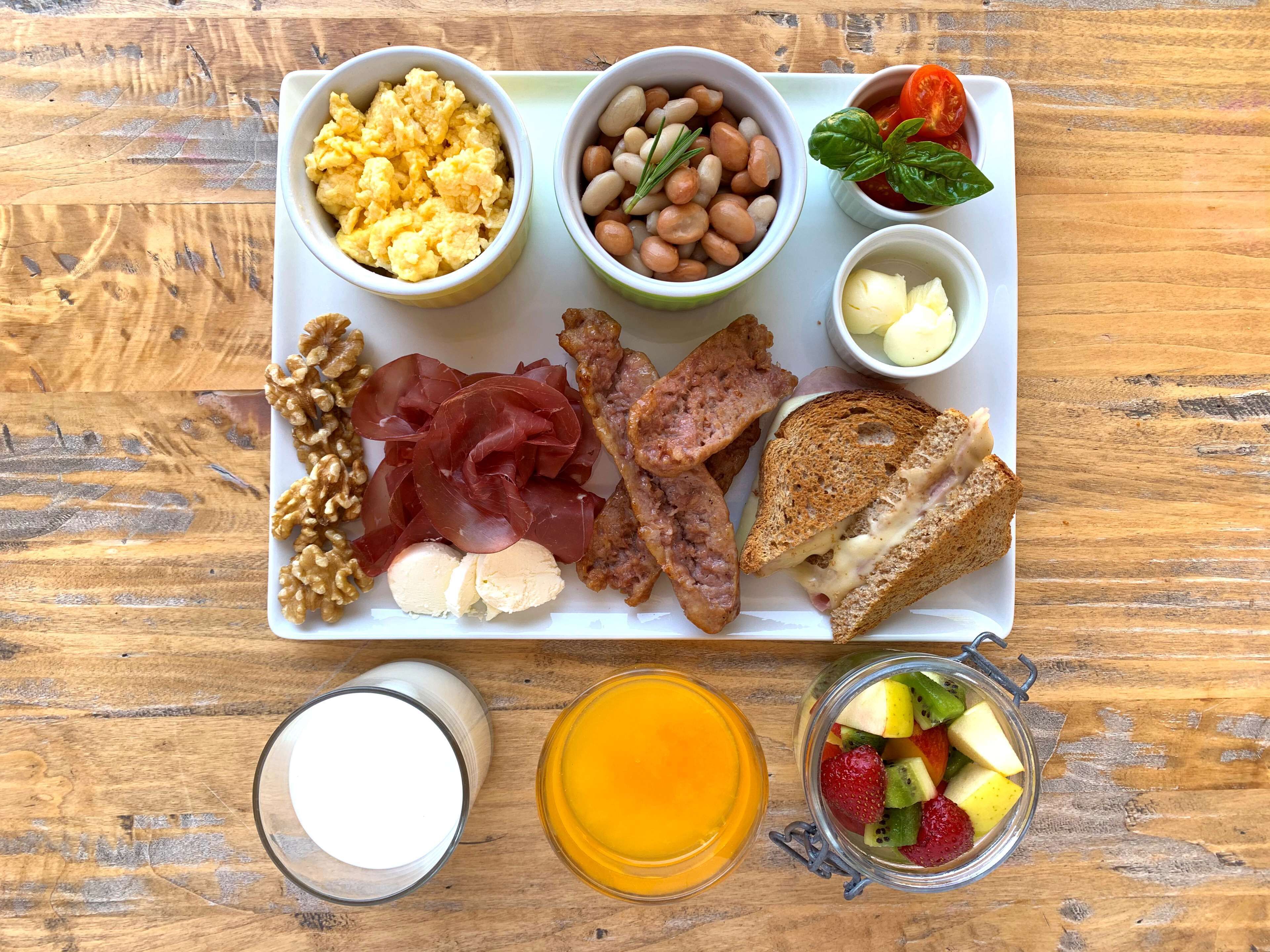 A breakfast tray with scrambled eggs, beans, tomatoes, basil, ham, cheese, toast, walnuts, prosciutto, and a fruit bowl.