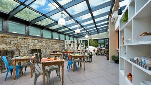 Un espace intérieur spacieux, aménagé avec des tables et des chaises, un mur en pierre et un plafond en verre qui laisse entrer la lumière naturelle.