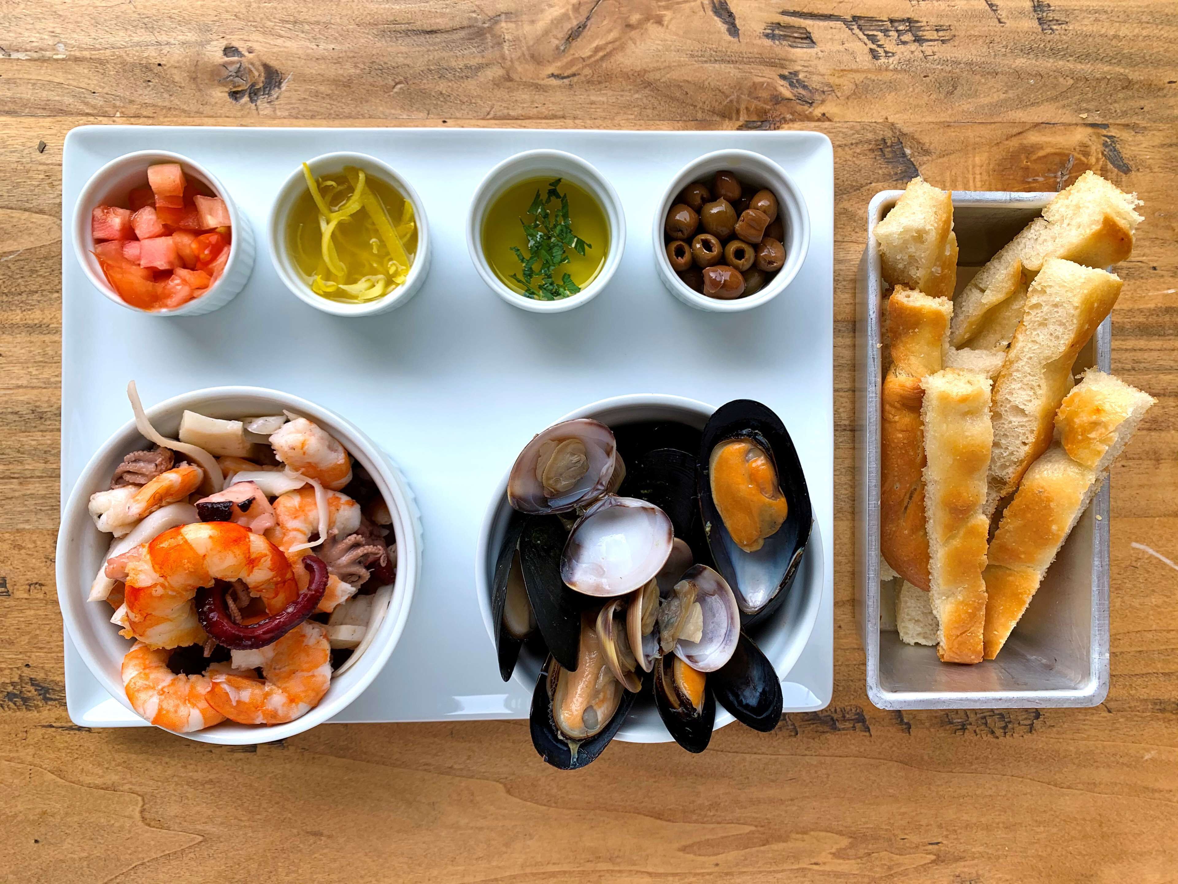 A seafood platter with shrimp, mussels, and clams, accompanied by small bowls of diced tomatoes, lemon slices, olives, and breadsticks.