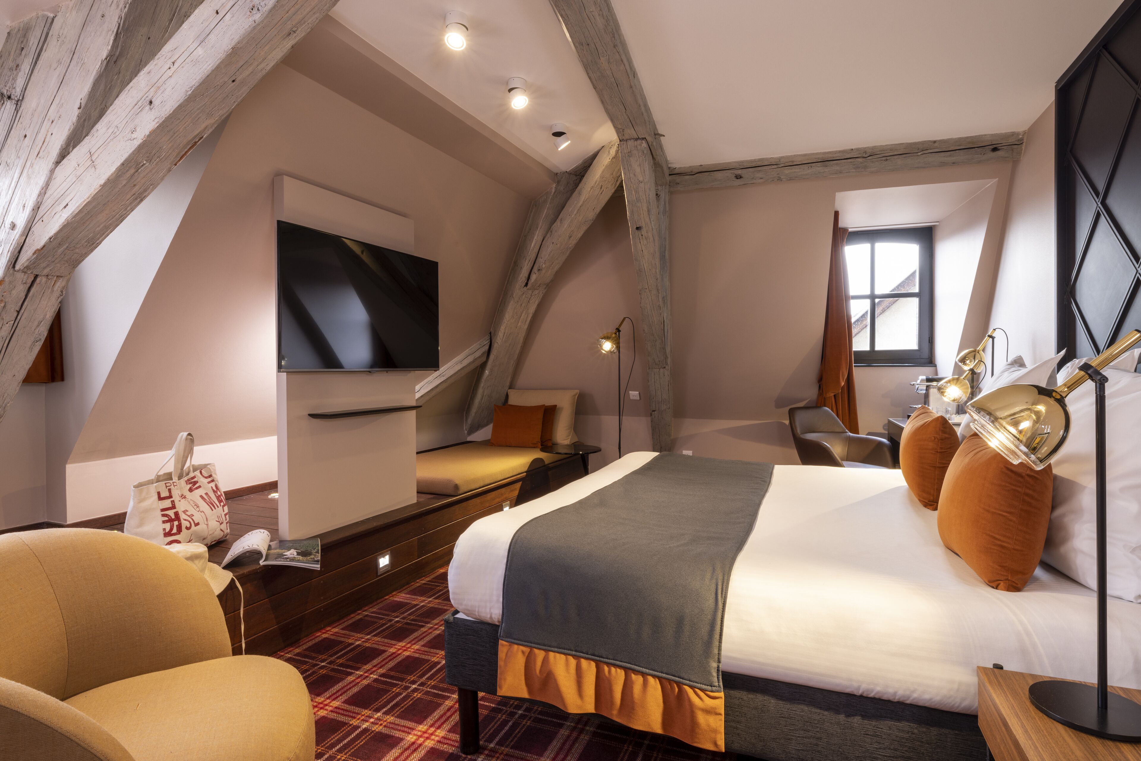 A modern hotel room with a large bed, a flat-screen TV, a desk with a lamp, and a window with curtains.