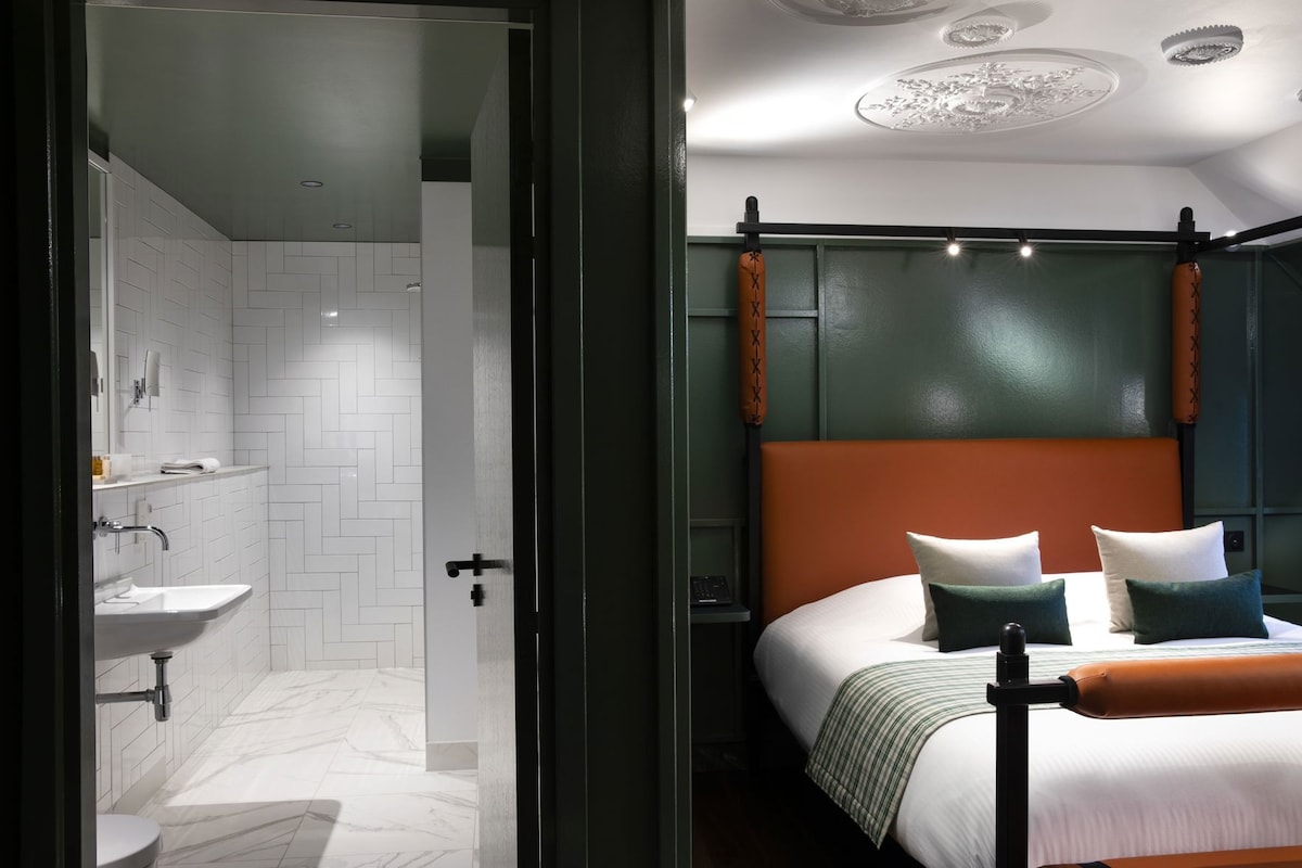Moderni hotellihuone, jossa on kylpyhuone, sänky tyynyineen ja yöpöytä.