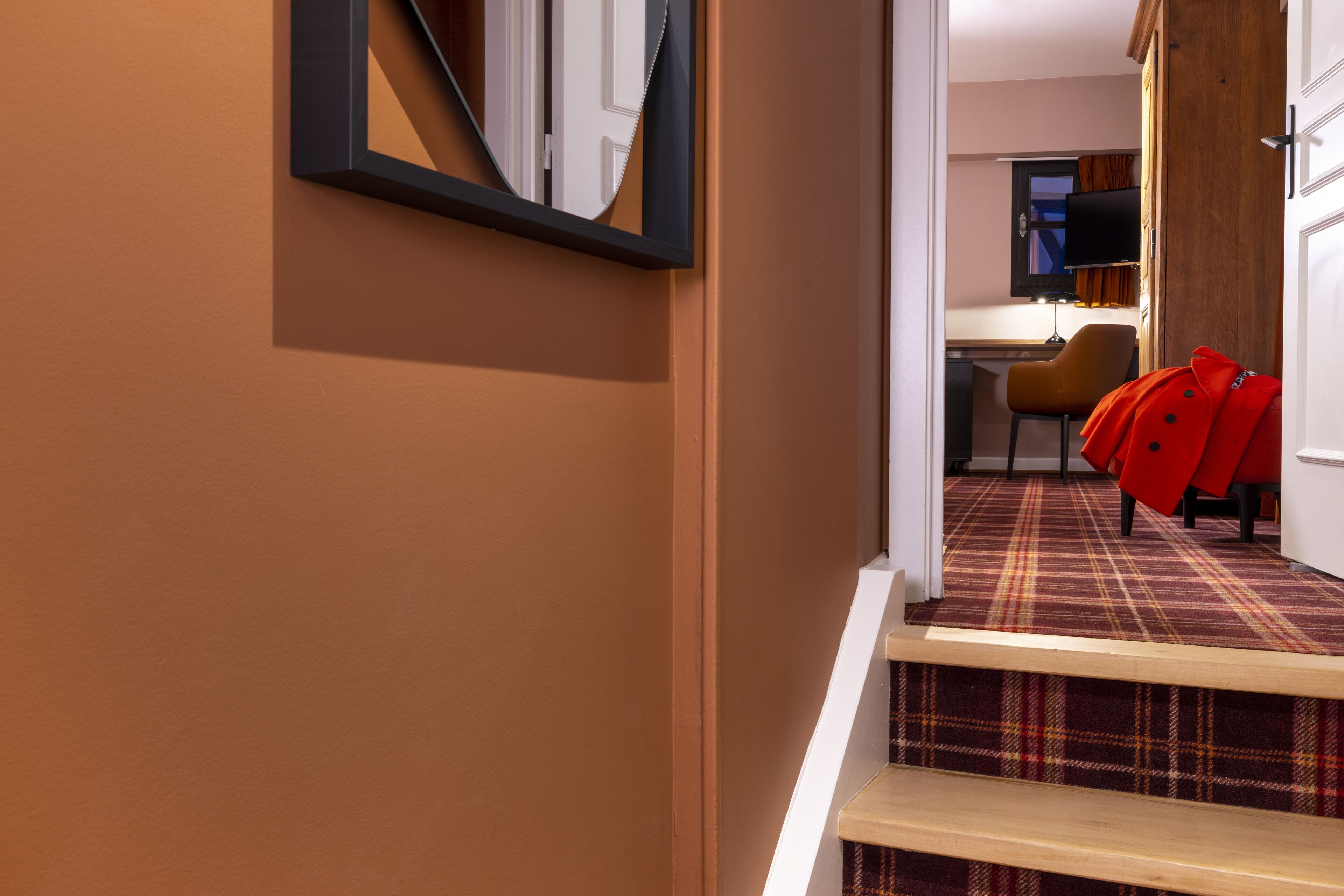 Un pasillo de hotel con alfombra escocesa, un banco de madera y un abrigo rojo colgado en la pared.