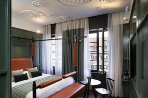 A modern hotel room with a large bed, a small table, a chair, and a window with curtains.