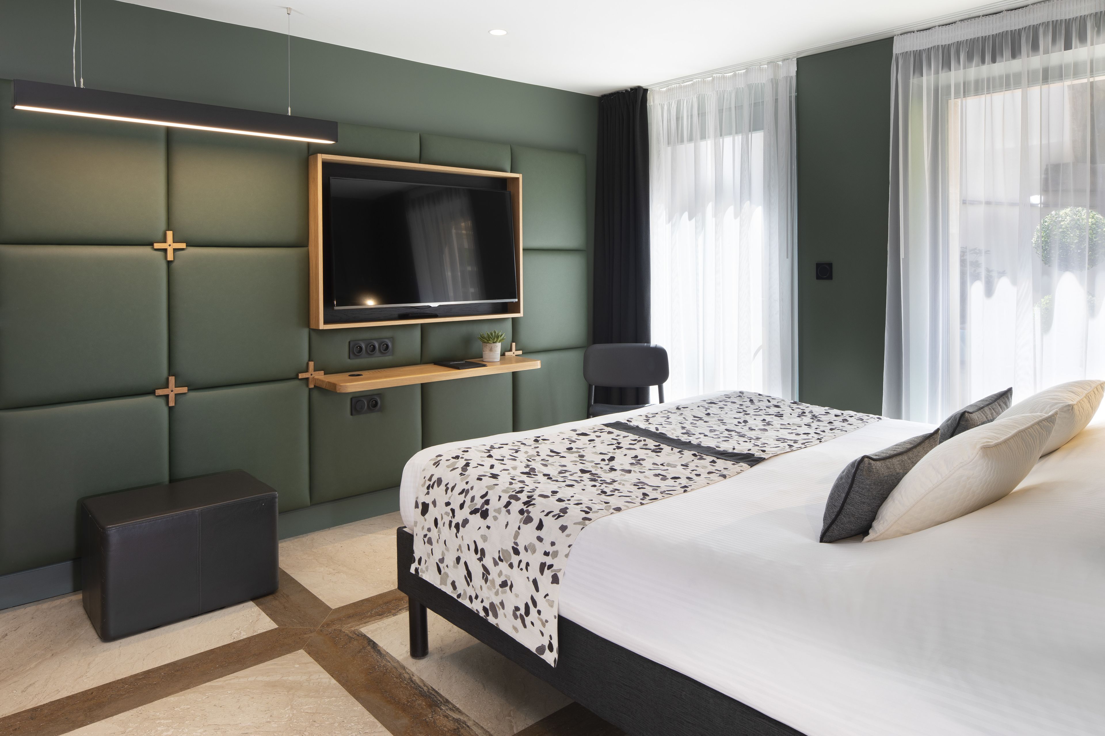 A modern hotel room with a large bed, a flat-screen TV mounted on a green padded wall, a small wooden side table, and a bench.