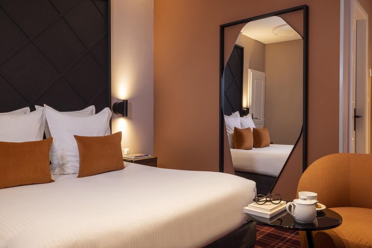 Hotellihuone, jossa on suuri sänky, yöpöytä, peili, tuoli ja pieni pöytä, jonka päällä on kuppi ja kirja.