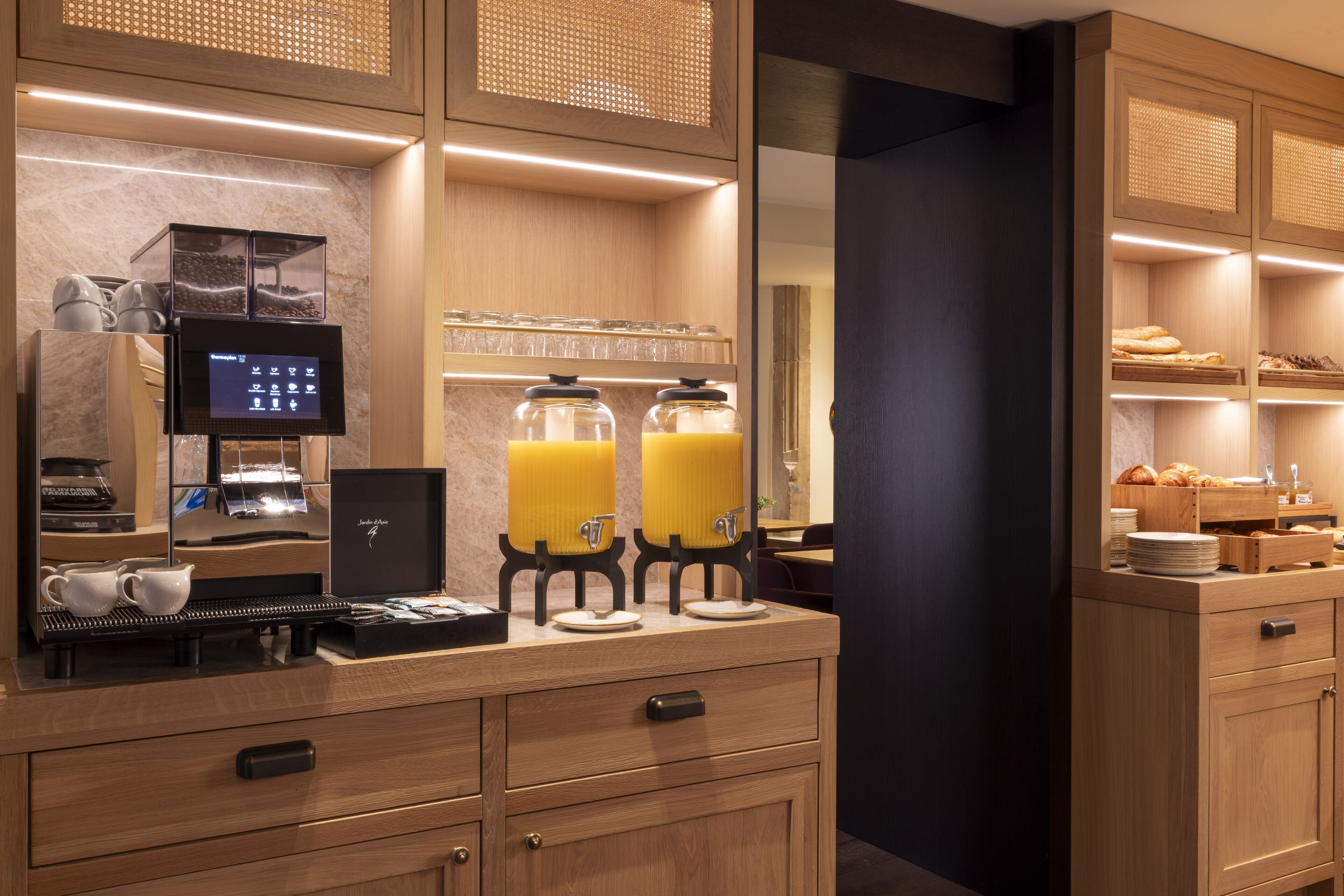 A modern hotel room with a wooden vanity, a coffee machine, a digital screen, and a breakfast bar with orange juice dispensers.