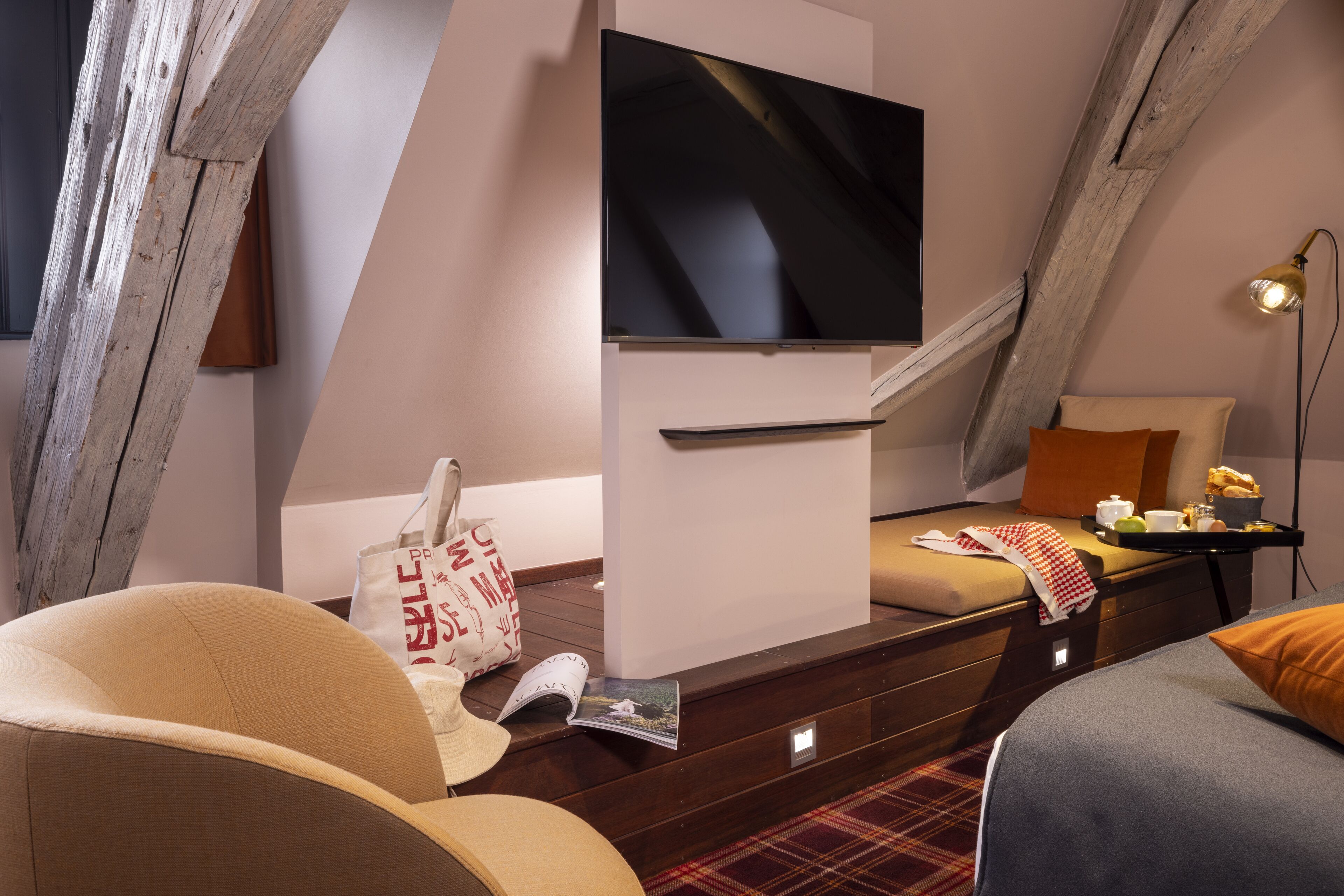 Una habitación de hotel moderna con un televisor de pantalla plana montado en la pared, una cama con manta escocesa, una mesita de noche con lámpara y una escalera de madera.