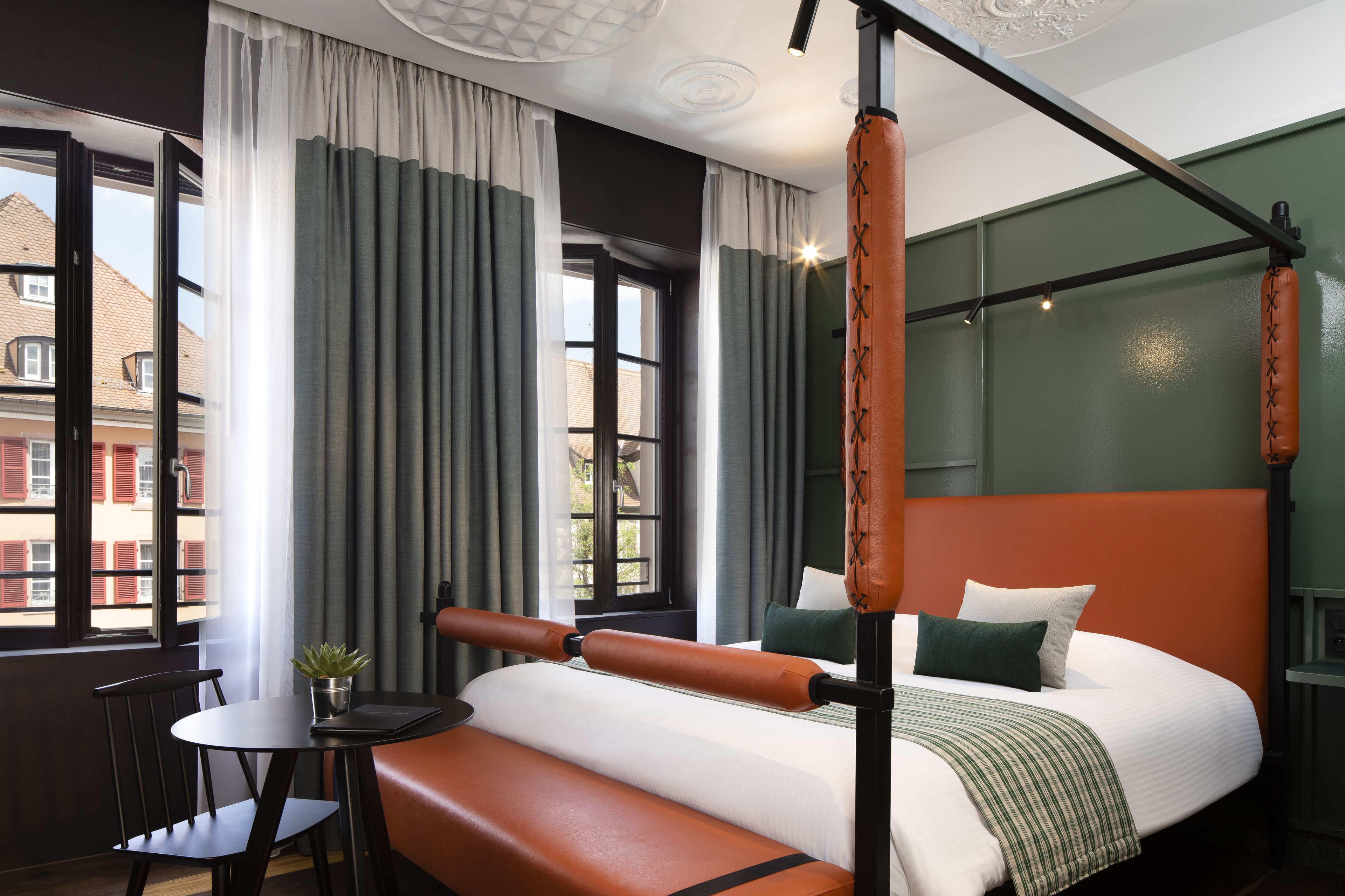 A modern hotel room with a large bed, a small table with a plant, and a view of buildings outside the window.