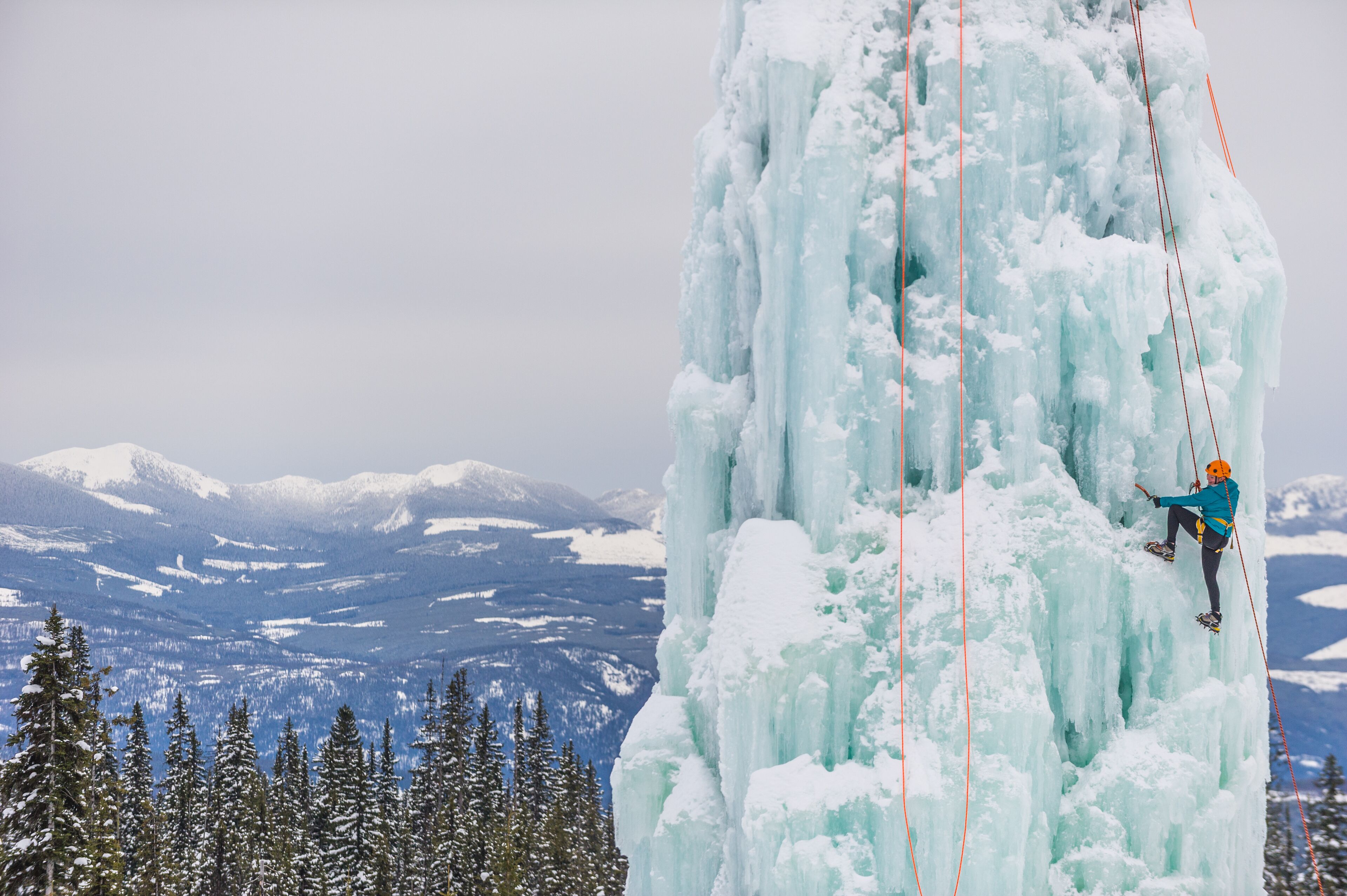 A person ice climbing a large ice formation with a rope, surrounded by snow-covered trees and mountains in the background.