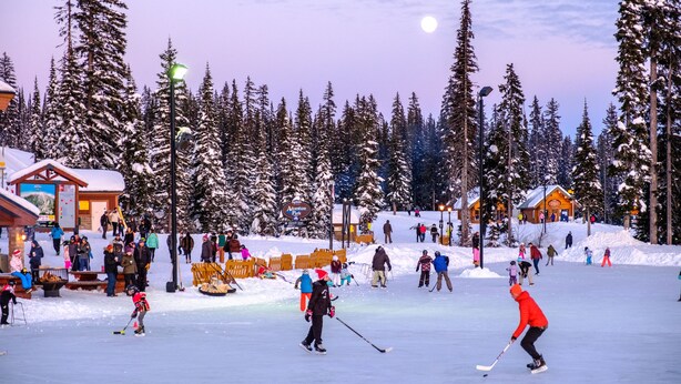 Un paisaje nevado al aire libre con gente esquiando y practicando snowboard, rodeado de altos pinos y cabañas de madera.