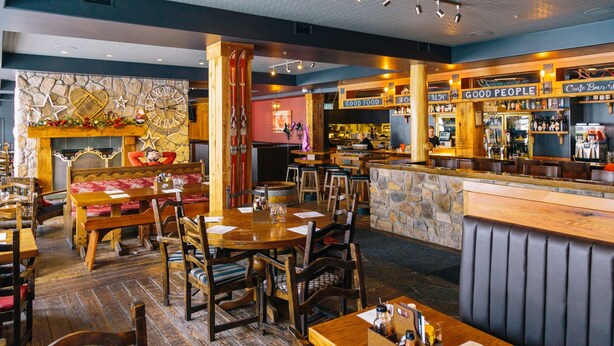 A cozy restaurant with wooden tables and benches, a stone fireplace, and a bar area with a sign reading "GOOD FOOD". - White Crystal Inn (Beaverdell)