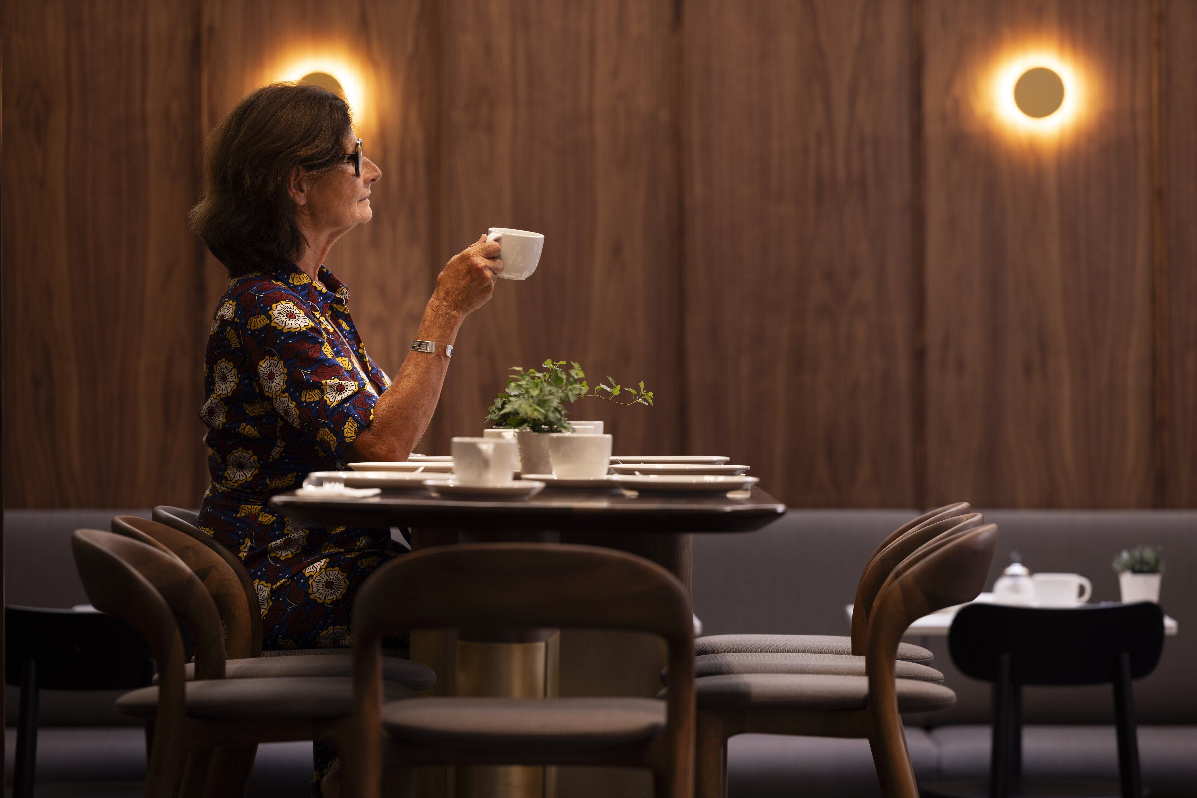 A person seated at a table with a cup, surrounded by chairs and a small plant.