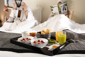 A hotel room with a bed, a tray with breakfast items, and two people reading magazines.