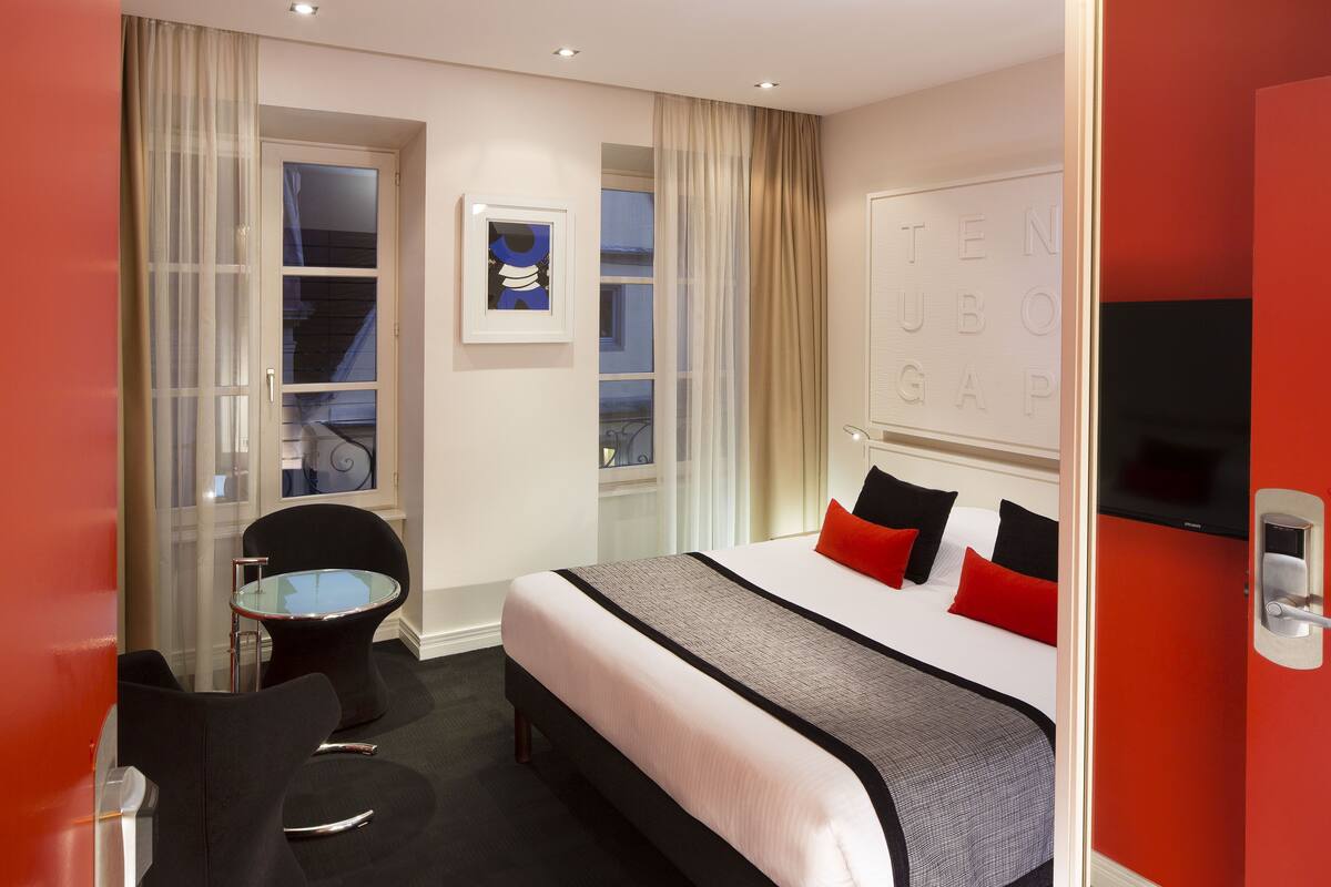 Habitación de hotel con cama, una mesita, dos sillas, televisor y ventana con cortinas.