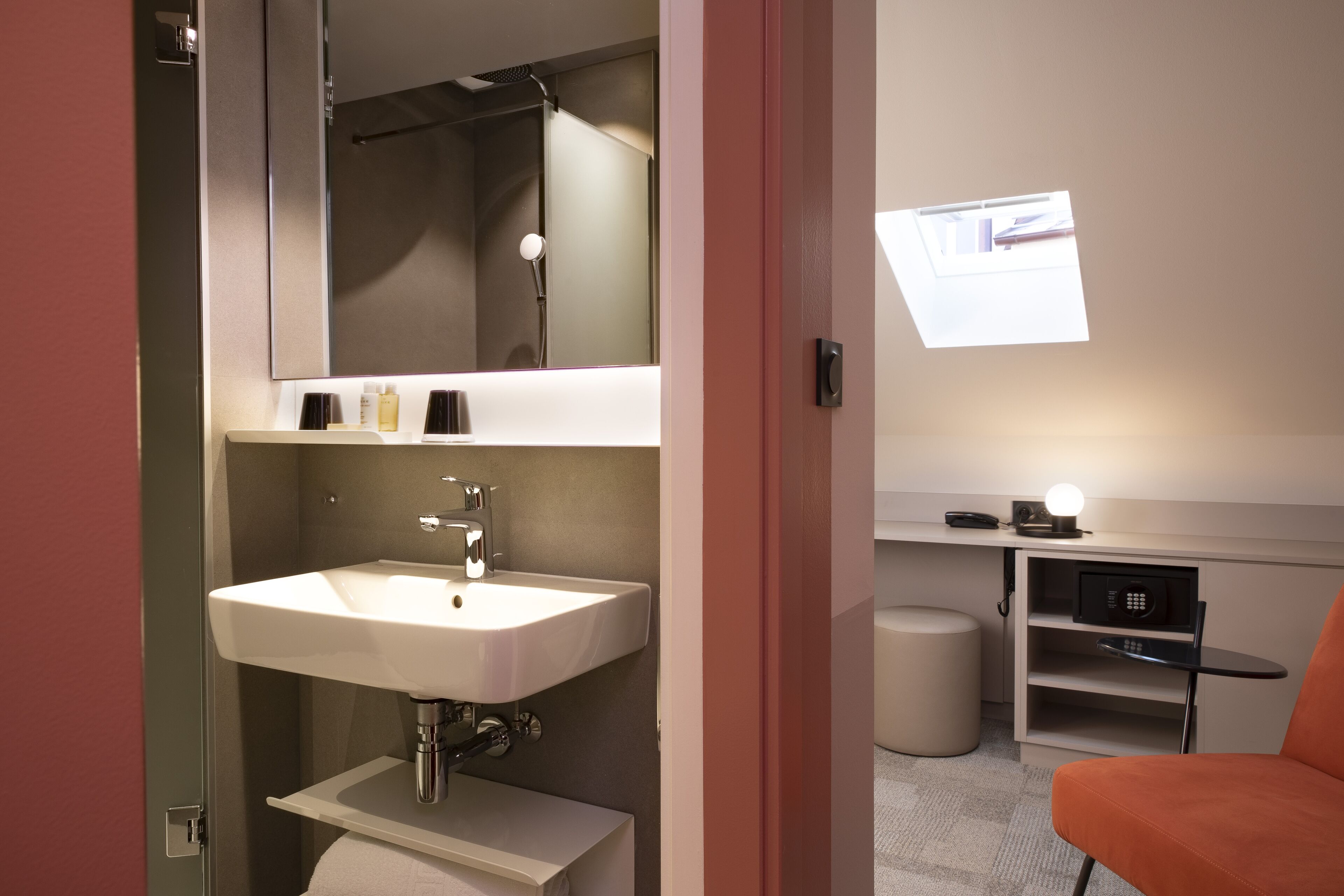 A modern bathroom with a sink, mirror, and shower. There is a small seating area with a chair and a table, and a window providing natural light.