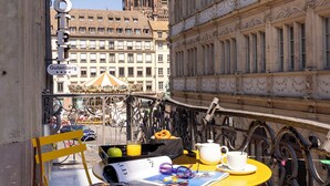 A yellow outdoor seating area with a table and chairs, a newspaper, and a drink. In the background, there is a large building with a tower and another building with a balcony. - Hôtel Gutenberg (Strasbourg)