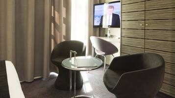 A hotel room with a modern design, featuring a flat-screen TV mounted on the wall, a glass-top table, and two contemporary chairs.