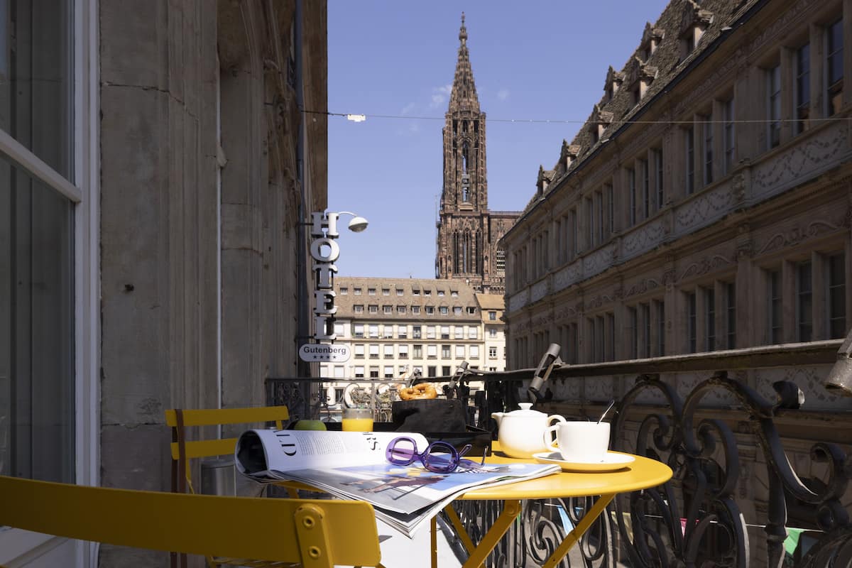 Un balcón con una mesa dispuesta para el desayuno, un periódico y una vista de una catedral al fondo.