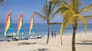 A beach with sailboats, palm trees, and a couple walking. - Hotel Riu Montego Bay - Adults Only - All Inclusive (Montego Bay)