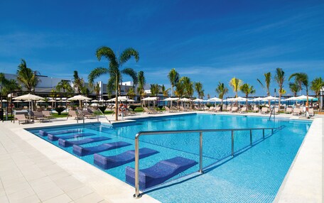 A large swimming pool with blue tiles, surrounded by lounge chairs and umbrellas, and palm trees in the background.