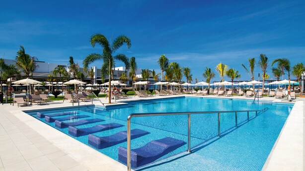 A large swimming pool with blue tiles, surrounded by lounge chairs and umbrellas, and palm trees in the background. - Hotel Riu Montego Bay - Adults Only - All Inclusive (Montego Bay)