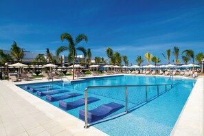 A large swimming pool with blue tiles, surrounded by lounge chairs and umbrellas, and palm trees in the background.