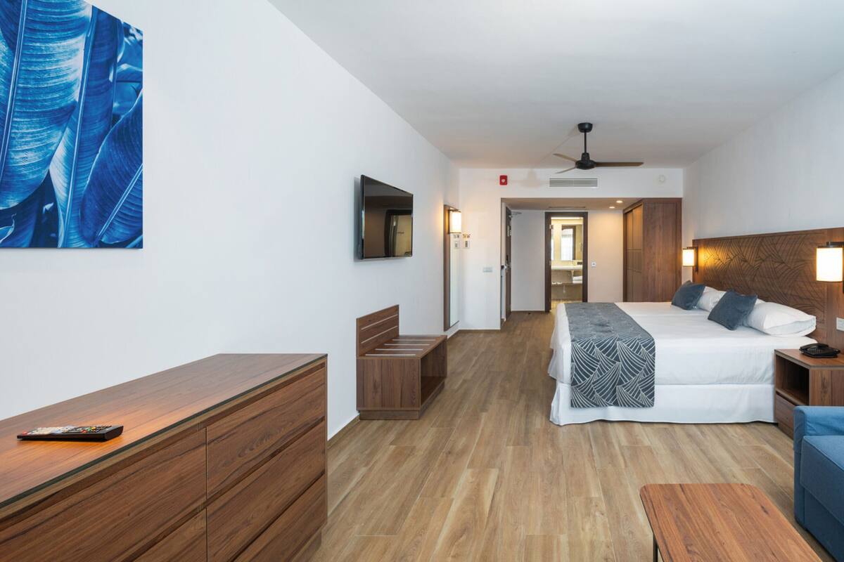Una habitación de hotel con cama, mesitas de noche, un sofá azul, suelo de madera y un ventilador de techo.
