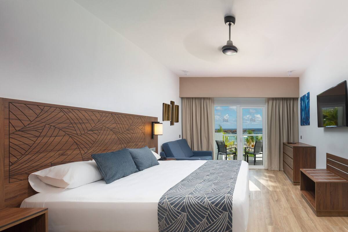 Una habitación de hotel moderna con una cama grande, cabecera de madera, un balcón con vistas y un televisor de pantalla plana.
